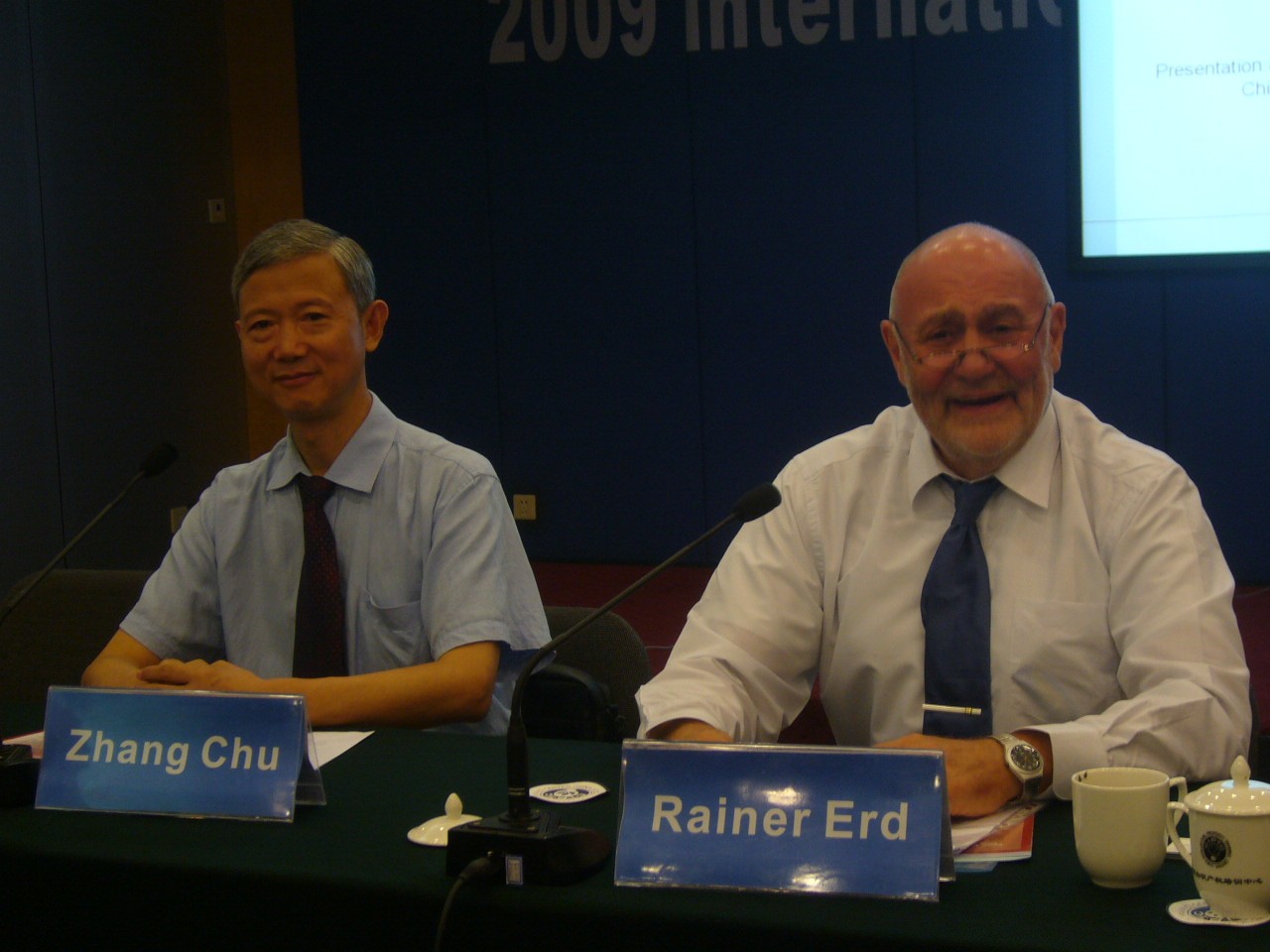 Zang Chu und Rainer Erd sitzen an einem Konferenztisch mit Mikrofonen