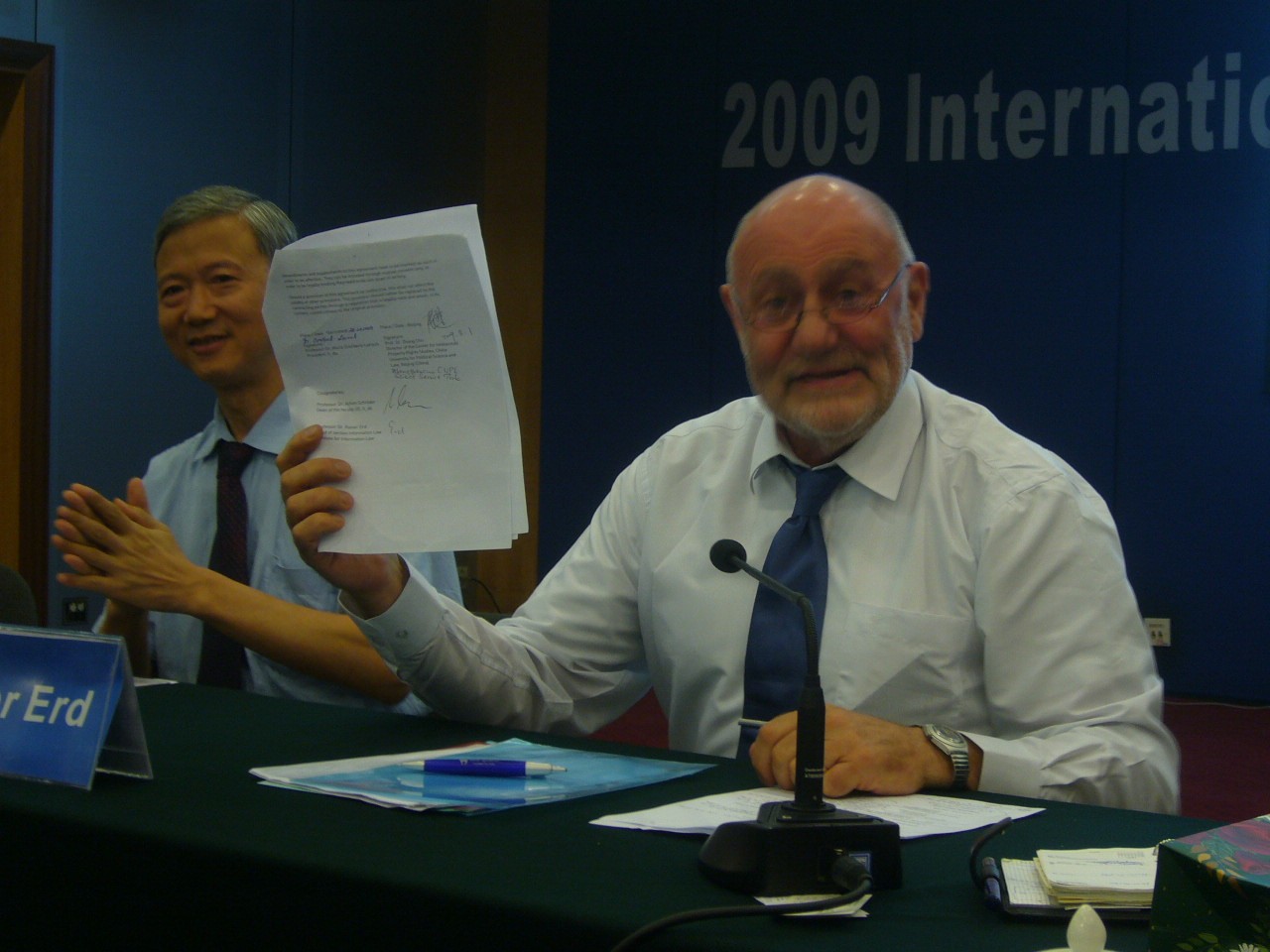Zwei Männer sitzen an einem Tisch, der eine Lacht und hält ein Dokument hoch, der andere klatscht und lächelt